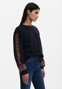 Felpa blu navy con motivi ricamati rossi e crema sulle maniche, con scollo rotondo e vestibilità comoda. Indossata con jeans di denim.
