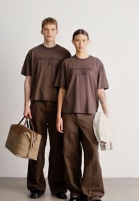 Brune bomuldst-shirts med logo, kombineret med matchende løstsiddende brune bukser. Den ene person holder en beige taske; den anden har en lys sweater.