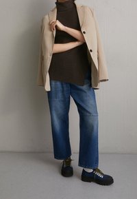 Beige Blazer, dunkler gerippter Rollkragenpullover, blaue weiter geschnittene Jeans und klobige blaue Schuhe mit schwarzen Sohlen und gelben Akzenten. Schlichte Texturen und Farben.