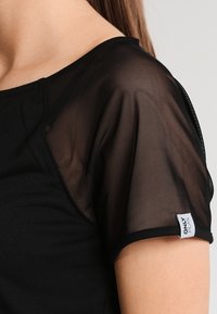 Svart topp med genomskinliga, lätta ärmar. Tyget är slätt med en mjuk textur. En liten vit etikett är sydd på ärmen.