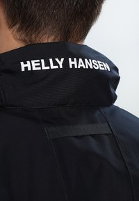 Crna jakna s visokim ovratnikom, koja sadrži bijeli logo "HELLY HANSEN". Ima glatku teksturu i minimalističke dizajnerske detalje.