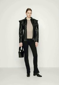 Casaco de pele sintética preto com ombros estruturados, top bege, jeans pretos em flare e uma pequena mala preta. Modelo a usar botas de bico.