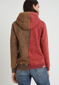 Bluza z kapturem o rozdzielonym wzorze; jedna strona czerwona, a druga brązowa. Wykonana z miękkiego materiału o fakturowanym wykończeniu oraz ściągaczami na mankietach.