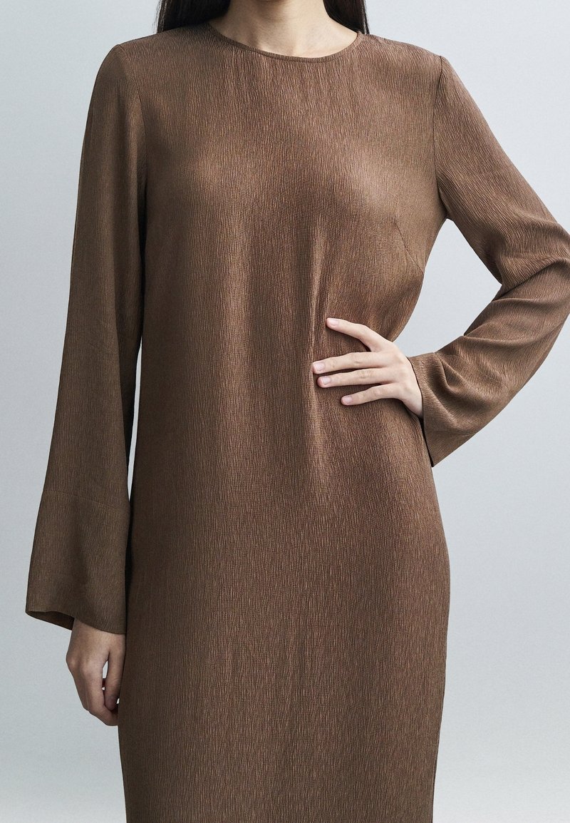 Femme portant une robe marron texturée à manches longues, une main posée sur la hanche, sur un fond clair uni.