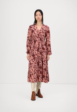 Long pink floral dress with long sleeves, a V-neck, and tiered ruffles. Features dark red flowers and a relaxed fit. Worn with brown ankle boots.