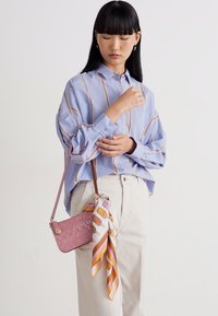 Nainen säätelee hihojaan, päällään vaaleansinihin raidoitettua paitaa, kermaisia housuja, kantaa vaaleanpunaista olkalaukkua, jossa on kuvioitu huivi yksinkertaista taustaa vasten.