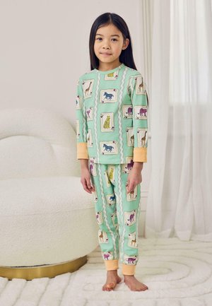 Young girl standing barefoot in mint green pajamas with colorful animal playing card prints in a light, cozy room.