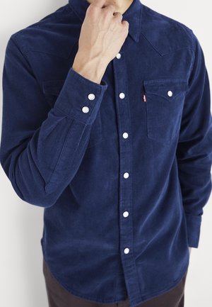 Mann in dunkelblauem Cordhemd mit weißen Knöpfen, Hand berührt das Kinn, Oberkörper sichtbar vor schlicht weißem Hintergrund.