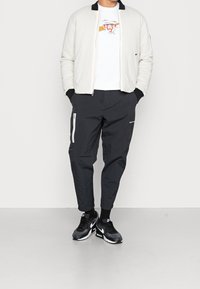 Herrbomberjacka i vitt med svart krage, ihop med svarta smalbyxor med sidofickor med dragkedja och svarta sportskor.