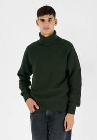 Maglione a collo alto lavorato a maglia di un verde scuro con texture a costine, vestibilità comoda e maniche lunghe, abbinato a jeans scuri.
