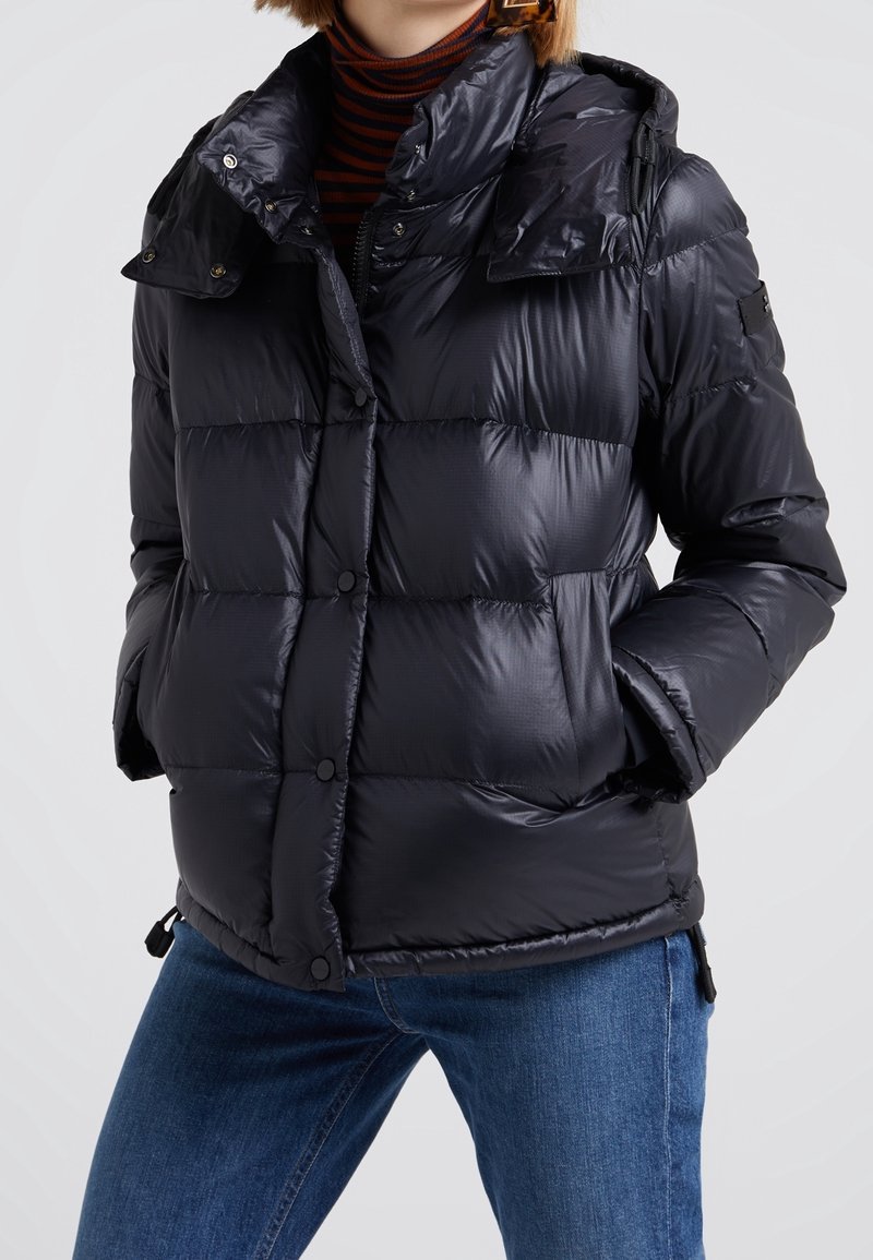 Person iført en sort quiltet dunjakke med hænderne i lommerne, kombineret med blå jeans og en sort og brun stribet rullekrave.