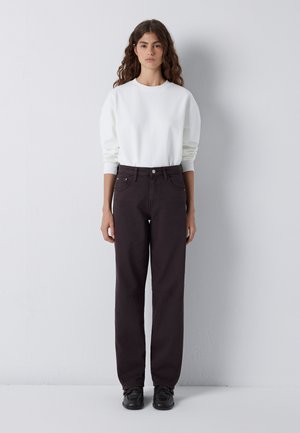 Woman with long curly hair stands against white wall wearing a white sweatshirt, dark straight-leg pants, and black shoes.