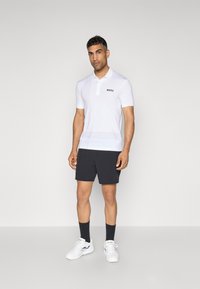 Weißes Poloshirt mit kurzen Ärmeln, gestreiftem Muster und schwarzem "BOSS"-Logo. Kombiniert mit dunklen Shorts und weißen Sportschuhen, getragen mit dunklen Socken.