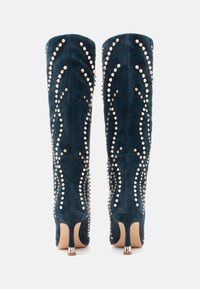 Augsti papēži līdz ceļiem, izgatavoti no tumši zaļa zamša, ar metāla studs, kas sakārtoti viļņveidīgā rakstā gar sāniem, un šauru papēdi.