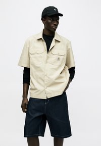 Άνδρας με μαύρο καπέλο, μπεζ πουκάμισο με κοντά μανίκια και κουμπιά πάνω από μαύρα μακριά μανίκια, και σκούρα τζιν σορτς, στέκεται με το ένα χέρι στην τσέπη.