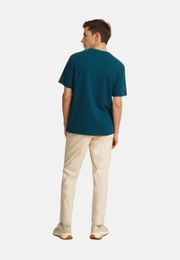 Türkiz rövid ujjú póló hátulról nézve, krém színű nadrággal és bézs tornacipővel, egy sima fehér háttér előtt.