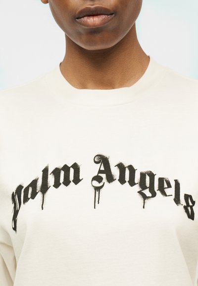 Detailný záber na osobu v bielej košeli s čiernym gotickým nápisom "Palm Angels" na hrudi, so šmuhovým efektom farby stekajúcej po texte.