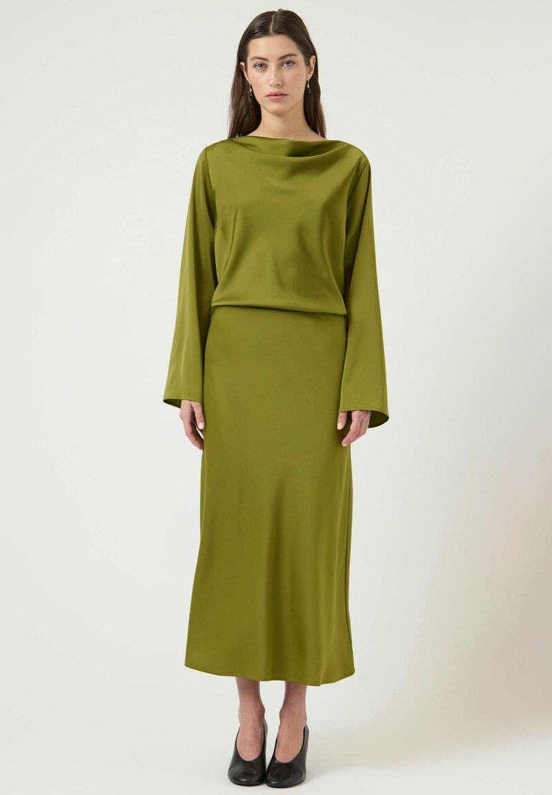 Vrouw met lang donker haar draagt een satijnen olijfgroene blouse met lange mouwen en een bijpassende midi-rok, gecombineerd met zwarte schoenen met hakken, staand tegen een witte achtergrond.
