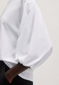 Witte, losse top met opstaande mouwen en een ronde halslijn. Zacht katoenen materiaal met een gladde textuur. Draag het met zwarte broeken.