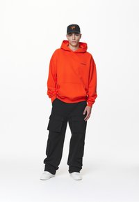 Tânăr stând, îmbrăcat într-un hanorac portocalie, pantaloni cargo negri, adidași albi și o șapcă neagră cu logo portocalie, pe un fundal alb.