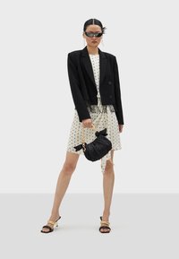 Blazer corto negro sobre un vestido blanco con lunares negros, sosteniendo un bolso negro texturizado con una cadena de oro y usando gafas de sol oscuras.