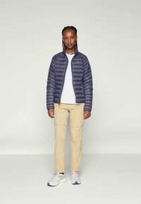 Moteris stovi vilkėdama tamsiai mėlyną pūkinę striukę, baltą marškinėlį, bege spalvos kelnes ir baltais sportiniais batais prieš lengvą, vienodą foną.