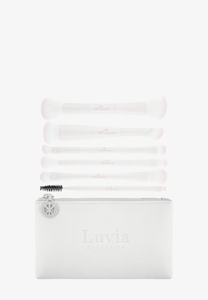 Six pinceaux de maquillage blancs avec des accents roses disposés au-dessus d'une pochette blanche zippée portant l'étiquette "Luvia Cosmetica".