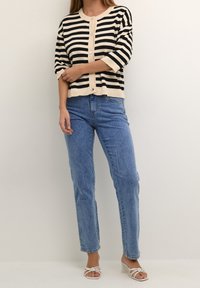 Schwarz-weiße gestreifte Strickjacke mit rundem Ausschnitt und Knopfverschluss, kombiniert mit hellblauen geraden Jeans und weißen Sandalen.