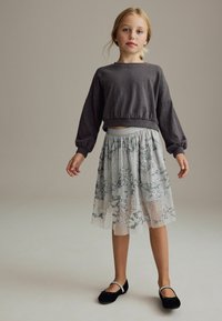 Sweat-shirt gris court avec des manches volumineuses, associé à une jupe grise transparente ornée de broderies florales argentées. Chaussures plates noires.