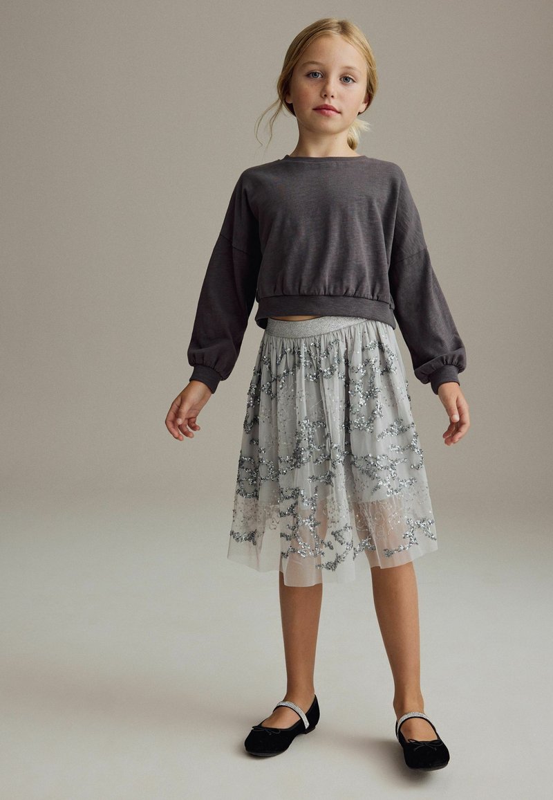 Sweat-shirt gris court avec des manches volumineuses, associé à une jupe grise transparente ornée de broderies florales argentées. Chaussures plates noires.