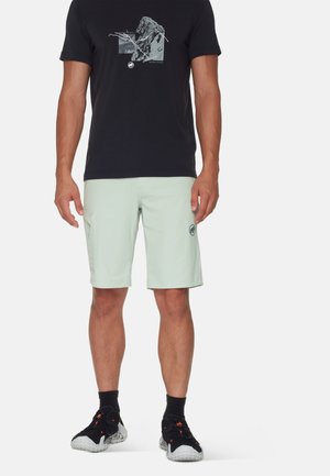 Man draagt een zwart graphic t-shirt, lichtgrijze shorts met zijzak, zwarte sokken en zwarte sportschoenen, staand tegen een witte achtergrond.