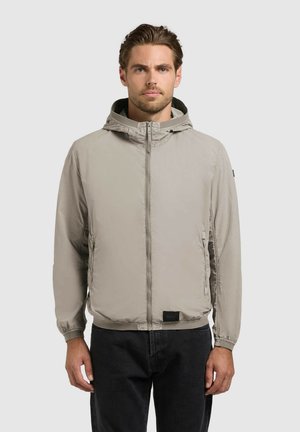 Hombre con chaqueta con capucha gris claro con cremallera y puños elásticos, y pantalones negros, de pie frente a un fondo liso y claro.