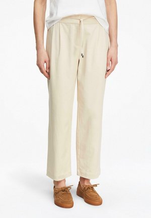 Personne portant un pantalon beige à jambes larges avec une taille à cordon de serrage et des chaussures décontractées marron à lacets, debout devant un fond blanc.