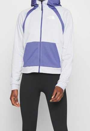 Person iført en hvid og lilla lynlåshætte med hætte og sorte leggings, der står mod en ensfarvet lys baggrund.
