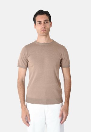 Uomo con capelli corti e scuri che indossa un maglione beige a maniche corte e pantaloni bianchi, in piedi davanti a uno sfondo chiaro uniforme.