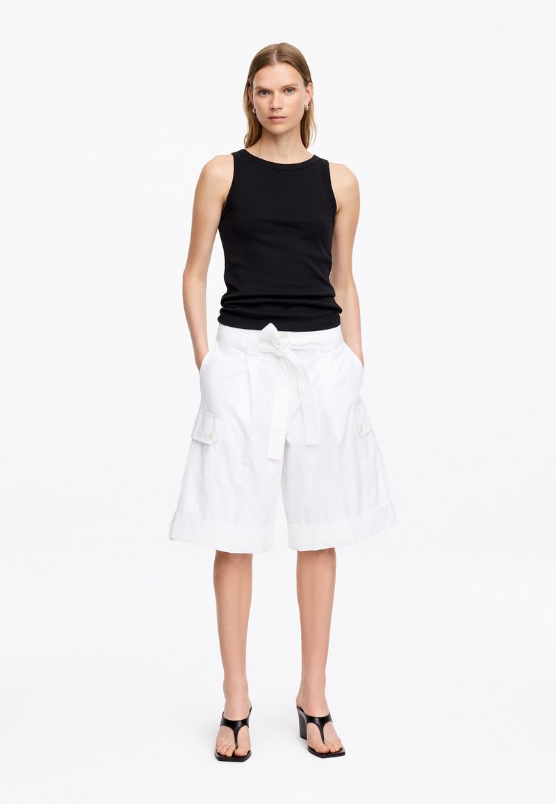 Femme debout portant un haut noir sans manches, un short cargo blanc jusqu'aux genoux avec ceinture, et des sandales noires à talons sur fond blanc uni.