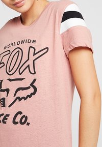 Rosa kortärmad T-shirt med svart grafiskt tryck, med vita och svarta randiga detaljer på axeln och en mjuk, texturerad tyg.