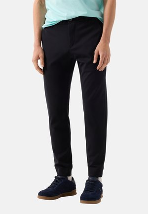 Mężczyzna noszący dopasowane czarne spodnie, jasnoniebieską koszulę, białe skarpetki i ciemnoniebieskie trampki, stojący na jednolitym tle.