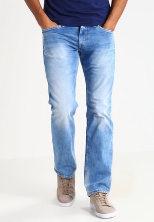 Mann trägt hellblaue ausgewaschene Jeans, Hände in den Taschen, und beige Sneaker, der auf einem schlichten weißen Hintergrund läuft.