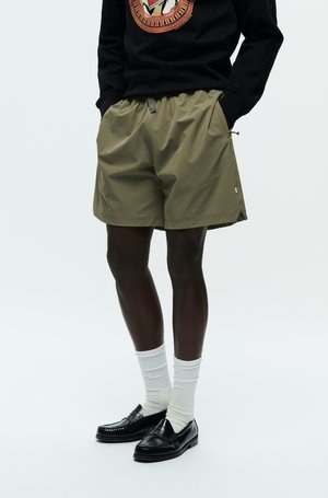 Olivgrüne Shorts mit einem elastischen Bund, seitlichen Taschen und einer lockeren Passform, kombiniert mit schwarzen Slippern und weißen gerippten Socken.