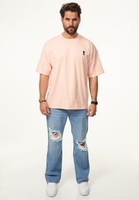 Rosa übergroßes T-Shirt mit einem kleinen Ananas-Logo, kombiniert mit hellblauen, verwaschenen Jeans mit großen Rissen und weißen Sneakers.