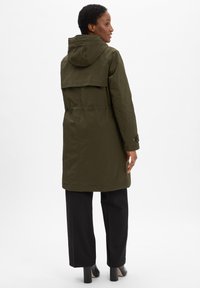 Woman wearing an olive green hooded long coat, black wide-leg trousers, and black heeled shoes, standing facing away with head turned right.