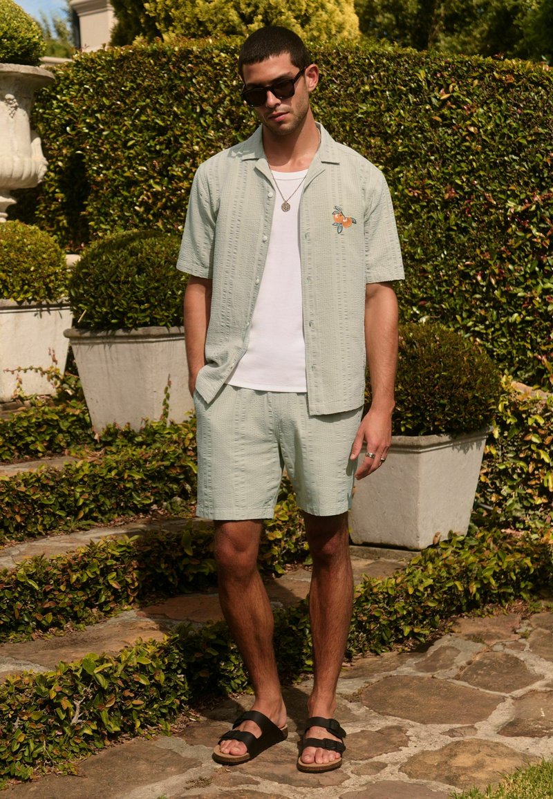 Mann trägt hellgrünes, kurzärmeliges Hemd mit Knöpfen und passende Shorts, weißes T-Shirt, schwarze Sandalen, Sonnenbrille, steht im Garten.