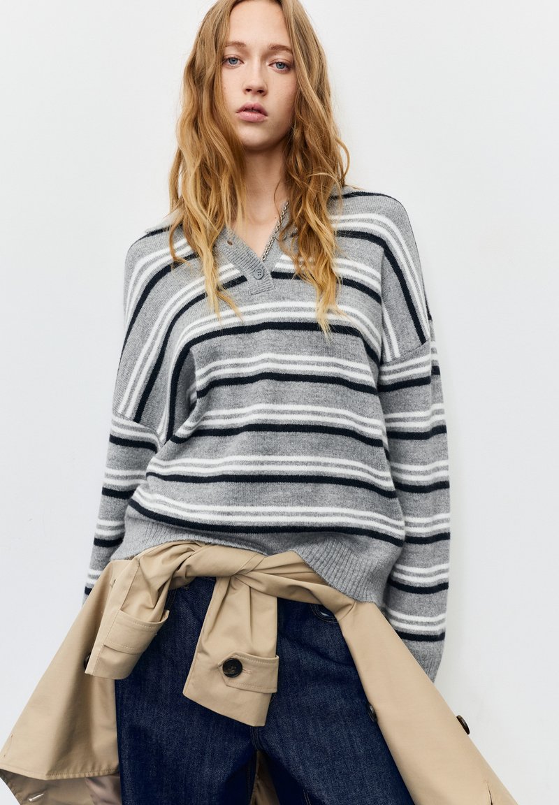 Szürke csíkos pulóver fekete-fehér vízszintes vonalakkal, V-nyakkivágással és bő fazonnal. Barna ballonkabát deréknál megkötve, sötét farmernadrág felett.