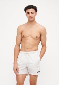 Pantaloni da bagno beige con vita regolabile tramite coulisse, tasche laterali e un'etichetta con logo nera. Tessuto morbido e taglio moderno, atletico.
