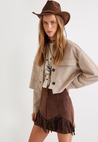 Cappello da cowboy in suede marrone, giacca cropped beige con due tasche frontali, t-shirt grafica color crema e gonna in faux-suede marrone con orlo frangiato.