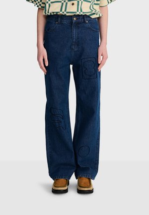 Personne portant un jean large bleu foncé avec des motifs cousus abstraits, associé à des chaussures marron et une chemise à motifs.