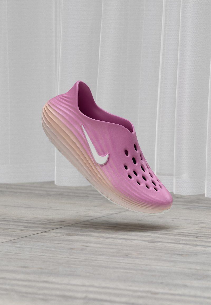 Ροζ παπούτσι slip-on με οπές και λευκό λογότυπο Nike, αιωρούμενο πάνω από ξύλινο πάτωμα, με φόντο λευκές κάθετες περσίδες.