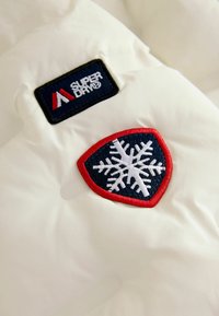Vitt vadderat jacka med en svart "Superdry"-patch och ett rött sköldemblem med en vit snöflingedesign. Slät, glänsande textur.