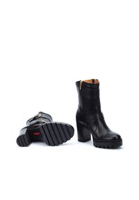 Botas de tobillo de cuero negro con un tacón robusto, cremallera lateral y detalles de correas horizontales. Suela de goma texturizada para un buen agarre.